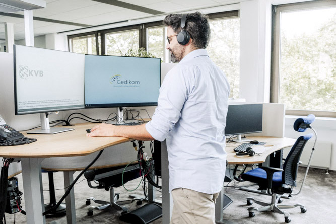 Ein Servicecenter Mitarbeiter der Gedikom arbeitet stehend an seinem Arbeitsplatz mit höhenverstellbarem Schreibtisch und zwei Monitoren.