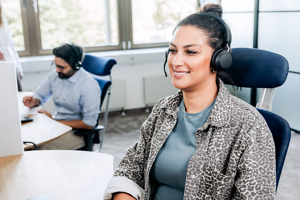 Servicecenter Agent / Agentin mit medizinischer Ausbildung Im Vordergrund: Freundlich lächelnde Gedikom Mitarbeiterin mit Headset im Bereich Servicecenter. Im Hintergrund: weiterer Servicenter Mitarbeiter.