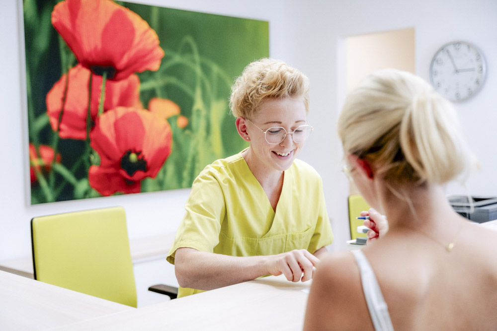 Freundlich lächelnde Mitarbeiterin der Gemeinschaftspraxis erklärt einer Patientin etwas am Empfang der Praxis.